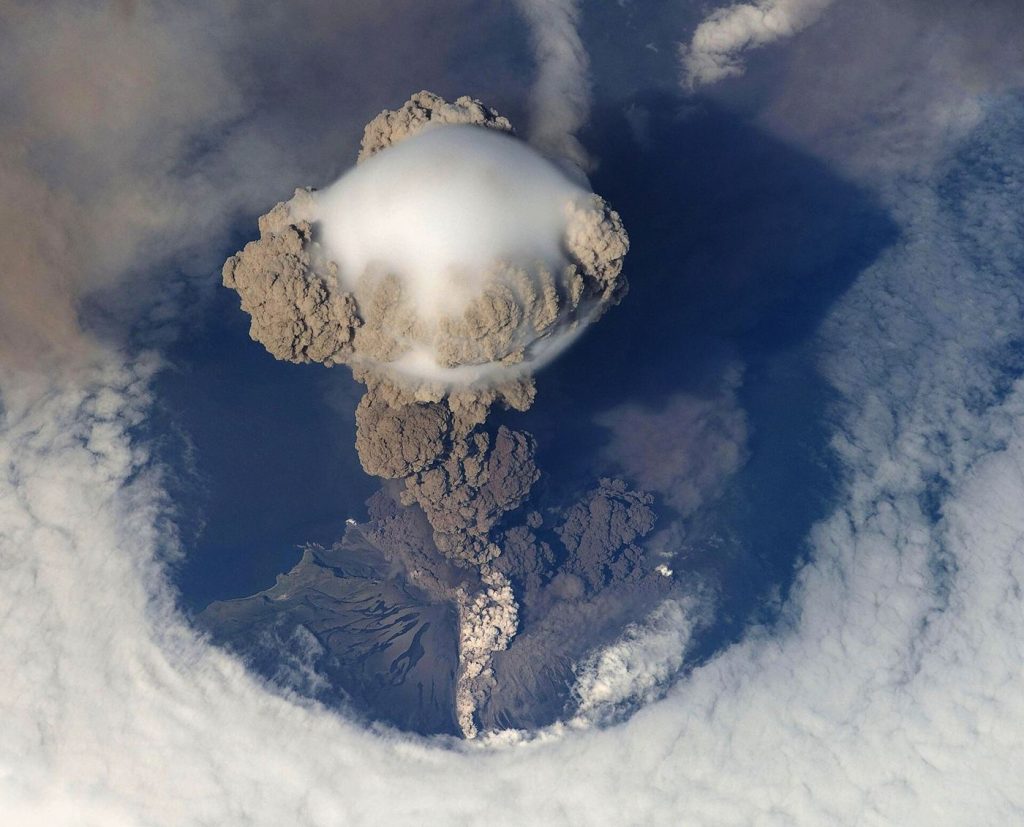 A volcanic eruption on an island sending a thick grey ash cloud into the sky above the coast.